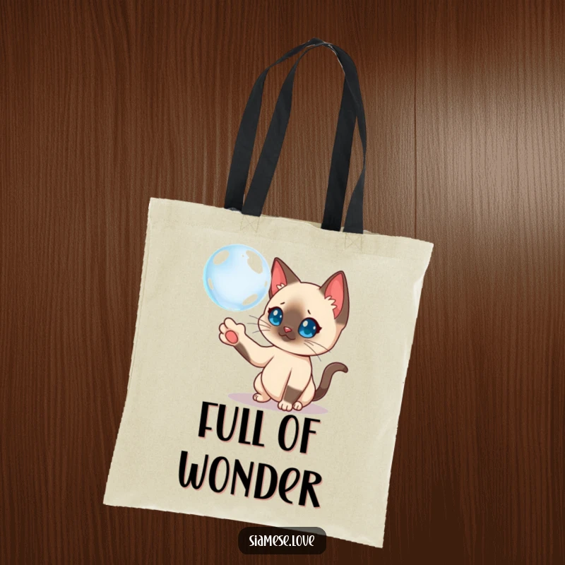 Funny Siamese cat batting at a floating bubble on a tote bag, highlighting playful curiosity.